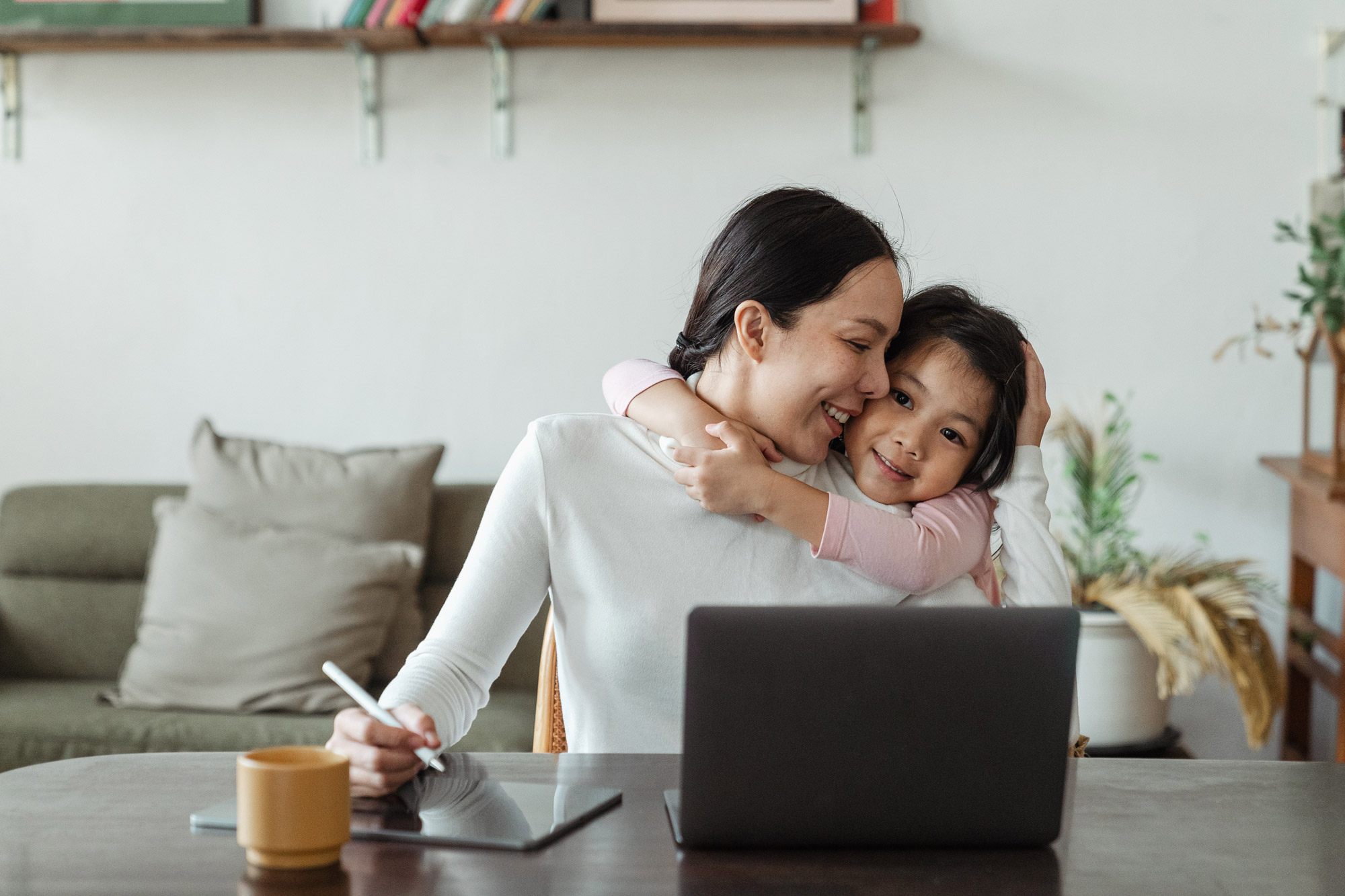 A small child wraps their arms around their mother while she is working on a tablet and computer