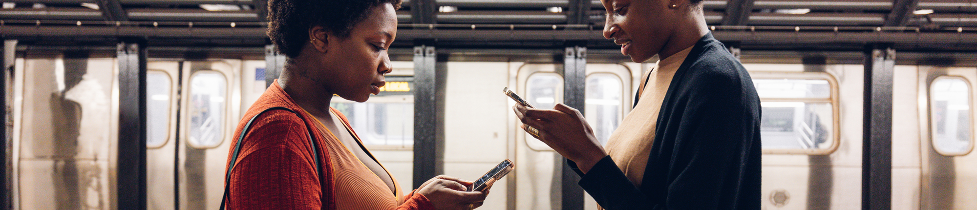 Two women look at their phones while waiting for a train to come in a subway station